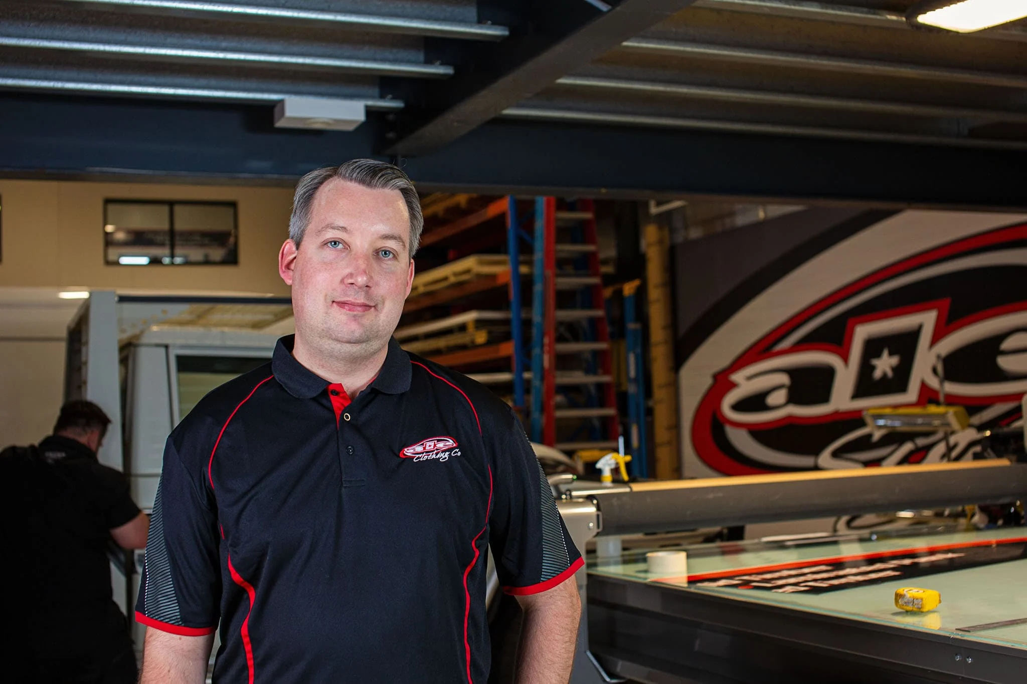 ALE team member in an embroidered polo in front of the ALE logo wall in the workshop